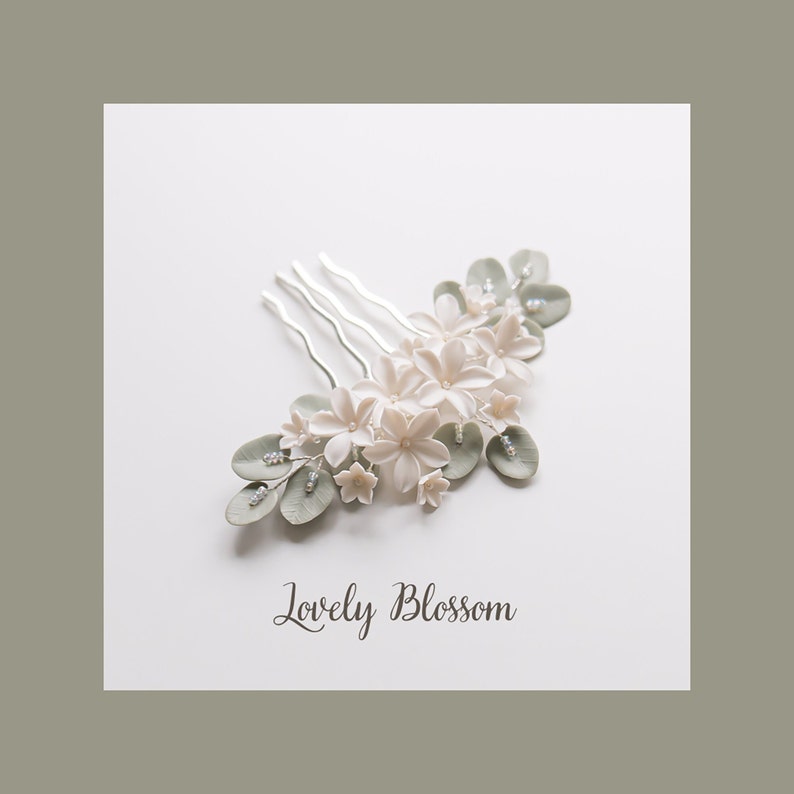 May include: A floral hair comb featuring white blossoms, pale green leaves, and small clear crystals. The comb has silver prongs and the text "Lovely Blossom" in script.