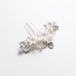 May include: A delicate floral hair comb featuring white flowers, pale green leaves, and small pearl accents. The comb has a silver metal base with three prongs. This elegant accessory is perfect for weddings or special occasions.