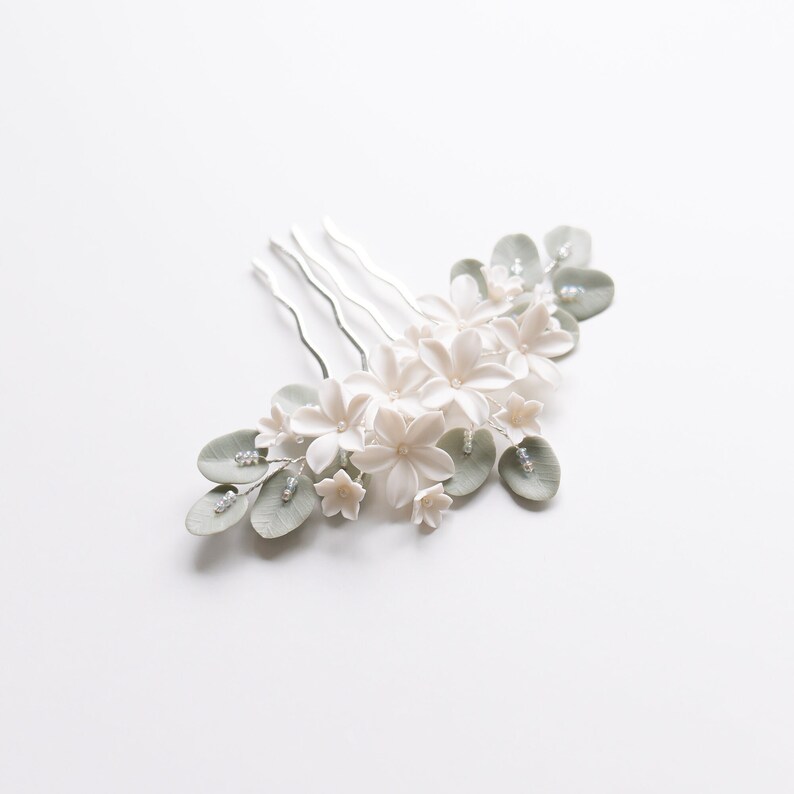 May include: A delicate floral hair comb featuring white flowers, pale green leaves, and small pearl accents. The comb has a silver metal base with three prongs. This elegant accessory is perfect for weddings or special occasions.