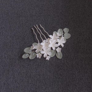 May include: A white floral hair comb featuring delicate, five-petal flowers and pale green leaves. The comb has three wavy prongs for securing in hair. The design is elegant and suitable for weddings or special occasions.