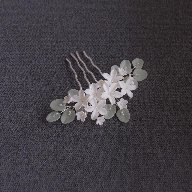 May include: A white floral hair comb featuring delicate, five-petal flowers and pale green leaves. The comb has three wavy prongs for securing in hair. The design is elegant and suitable for weddings or special occasions.