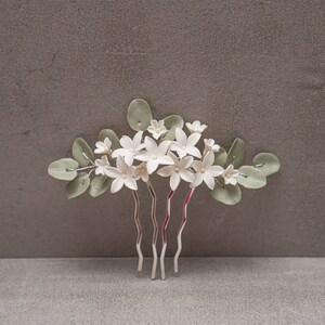 May include: A floral hair comb featuring white flowers with pearl centers and sage green leaves. The comb has silver-toned metal prongs. The flowers are arranged in a cluster, creating a delicate and elegant accessory.