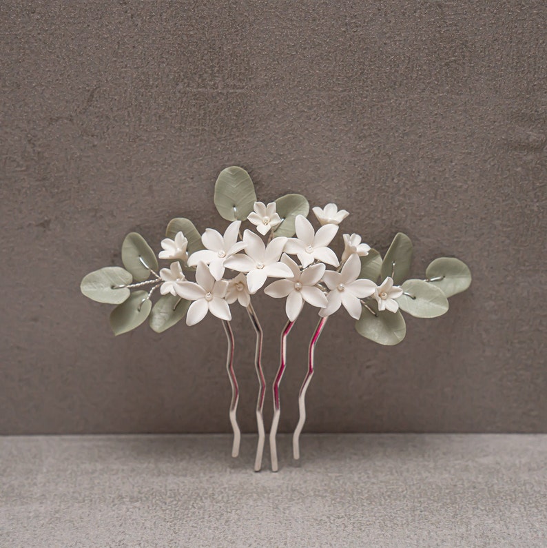 May include: A floral hair comb featuring white flowers with pearl centers and sage green leaves. The comb has silver-toned metal prongs. The flowers are arranged in a cluster, creating a delicate and elegant accessory.