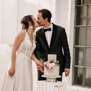 Könnte beinhalten: Eine zweistöckige Hochzeitstorte mit Blumendekoration, platziert auf einem weißen Ständer. Ein Paar küsst sich vor der Torte. Der Mann trägt einen schwarzen Smoking. Die Frau trägt ein weißes Brautkleid.