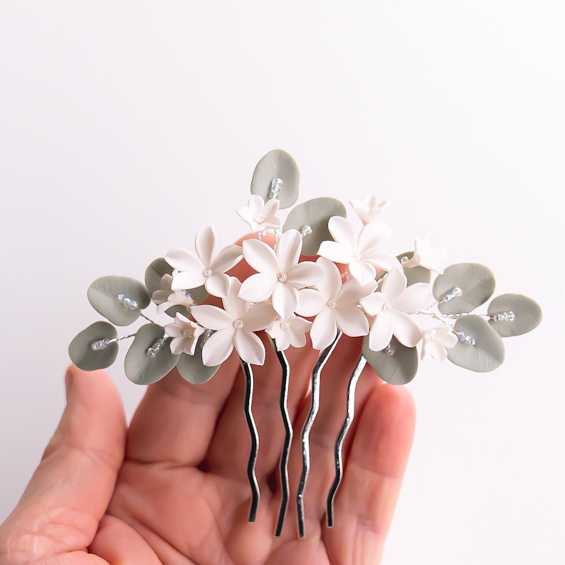 May include: A floral hair comb featuring white flowers, gray leaves, and silver accents. The comb has a silver base with multiple prongs. The flowers have small pearl-like centers, and the leaves are a muted green.