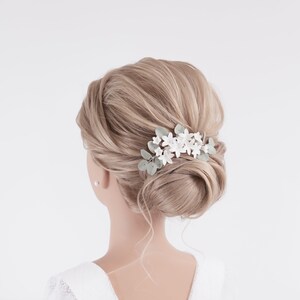May include: A bridal hair comb featuring white floral accents and delicate green leaves. The comb is set against a backdrop of styled blonde hair, creating an elegant and romantic look. Perfect for weddings or special occasions.