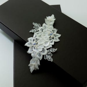 Könnte beinhalten: Ein weißes Spitzenstrumpfband mit einem komplizierten floralen Stickmuster und zarten Blattmotiven. Das Design enthält kleine, hellblaue Perlenakzente, die in der Mitte der weißen Blütenblätter sitzen und eine detaillierte Textur erzeugen.