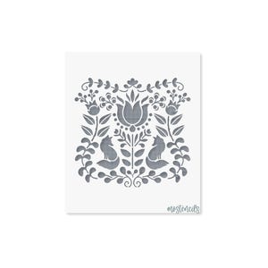Op de afbeelding: Een vierkant sjabloon met een symmetrisch grijs ontwerp van twee vossen die naar elkaar toe kijken, omringd door gestileerde bloemen, bladeren en bessen. Het woord mostencils staat in een cursief lettertype in de rechterbenedenhoek.