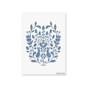 Può includere: Un foglio stencil rettangolare con un design di arte popolare blu e simmetrico. Il motivo mostra due conigli rivolti verso un fiore centrale, circondati da foglie, steli e boccioli floreali stilizzati. Il testo "mostencils" è stampato in basso a destra.