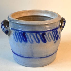 Könnte beinhalten: Ein hellblaues Keramikgefäß mit einem dunkelblauen Wellenmuster. Der Topf hat zwei Griffe und einen hellbraunen Rand. Das blaue Design ist eine Reihe von vertikalen und horizontalen Wellenlinien. Der Topf wird wahrscheinlich für dekorative oder funktionelle Zwecke verwendet.