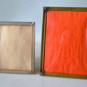 Op de afbeelding: Twee vintage fotolijsten. Eén is rechthoekig met een zilverkleurig metalen frame en een getextureerd, lichtbruin interieur. De andere is een grotere rechthoekige lijst met een goudkleurig metalen frame en een oranje interieur.