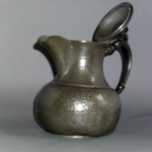 May include: A pewter pitcher with a hammered finish and a hinged lid. The pitcher has a rounded body and a curved handle.