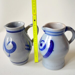 Può includere: Due brocche in ceramica grigia con disegni a spirale blu e manici. Le brocche sono alte circa 25 cm, con un beccuccio per versare. Le brocche sono affiancate, con un righello giallo in mezzo.