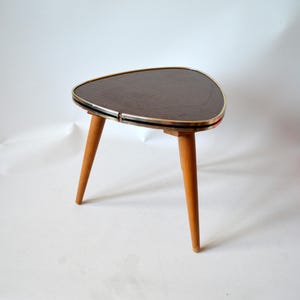 Op de afbeelding: Een driepotig bijzettafeltje met een donkerbruin, driehoekig blad met afgeronde hoeken. Het tafelblad is afgewerkt met een dunne metalen rand. De tafel staat op drie taps toelopende, lichtgekleurde houten poten die naar buiten staan.