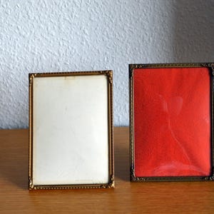 Puede incluir: Dos marcos de fotos rectangulares con bordes metálicos en tono dorado fino que presentan un detalle de borde con cuentas. Un marco contiene un inserto blanco liso, mientras que el otro marco tiene un inserto rojo vibrante con un patrón floral sutil.
