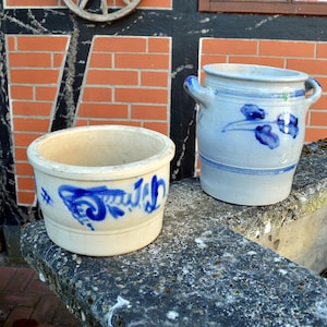 May include: Two antique stoneware pots with blue floral designs. One is a cream-colored, low-profile pot with a blue design and text. The other is a taller, blue-gray pot with handles and blue floral accents. Both are on a stone surface.