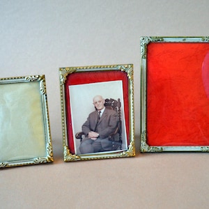 Puede incluir: Tres marcos de fotos antiguos con bordes dorados ornamentados. Un marco contiene una fotografía de un hombre con traje, mientras que otro tiene un respaldo rojo. El tercer marco tiene un respaldo de color crema.