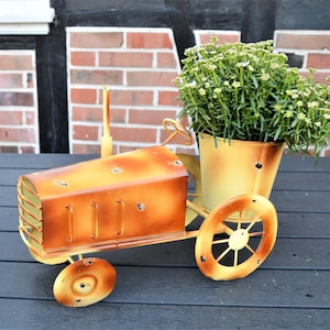 Könnte beinhalten: Ein rostiger orangefarbener Traktor aus Metall mit einem gelben Topf aus Metall, gefüllt mit weißen Blumen. Der Pflanzer hat auf jeder Seite ein Rad und ein kleines Rad vorne.