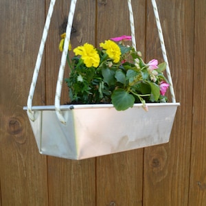 Könnte beinhalten: Ein weißer, hängender Blumentopf aus Metall mit gelben und rosa Blüten. Der Topf hängt an zwei weißen Schnüren an einer Holzoberfläche.