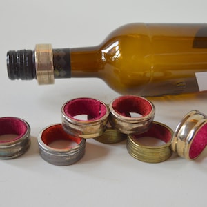 May include: A collection of wine bottle rings with a red velvet interior. The rings are made of metal in various finishes, including silver and gold. A brown wine bottle is in the background.