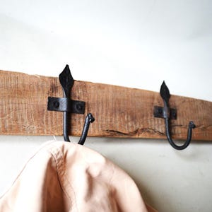 Könnte beinhalten: Ein rustikaler Holz-Kleiderständer mit drei schwarzen Metallhaken. Die Haken haben ein dekoratives, spitzes Design und sind mit Metallhalterungen am Holz befestigt. Eine hellbraune Jacke hängt an einem der Haken.
