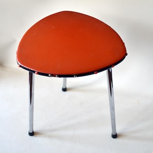 Könnte beinhalten: Ein orangefarbener Retro-Hocker mit dreieckiger Sitzfläche und verchromten Beinen. Der Sitz hat einen schwarzen Rand und kleine silberne Nieten. Der Hocker ist etwa 30 cm breit und hat drei Beine.