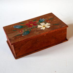 Op de afbeelding: Een kleine, rechthoekige houten doos met een scharnierend deksel. Het is roodbruin gebeitst. Het deksel is versierd met handgeschilderde bloemen in rood, wit en blauw. De doos wordt waarschijnlijk gebruikt voor het opbergen van kleine voorwerpen.