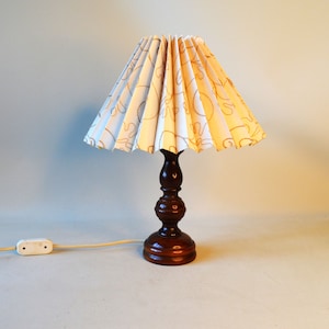 Könnte beinhalten: Eine Tischlampe aus Holz mit einem weißen, gefalteten Lampenschirm mit braunem Blumenmuster. Die Lampe hat ein braunes Kabel und einen weißen Schalter.