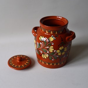 May include: A hand-painted terracotta jar with a lid. The jar has a floral design with yellow flowers, green leaves, and white accents. The lid has a small knob on top.