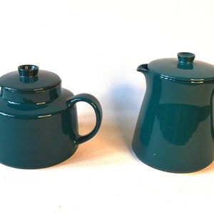 Pu&ograve; includere: Un set da t&egrave; in ceramica blu-verde composto da una teiera e un lattiera. La teiera ha un corpo rotondo e un coperchio con un pomo. La lattiera ha un corpo alto e sottile e un beccuccio.