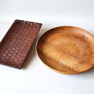 Puede incluir: Dos bandejas de servir de madera. Una es una bandeja rectangular con una superficie texturizada. La otra es una bandeja redonda con una superficie lisa.