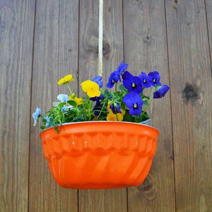 Könnte beinhalten: Ein leuchtend orangefarbener Hängepflanztopf mit gelben und blauen Blumen. Der Topf ist aus Metall und hat einen gewellten Rand.