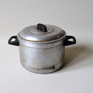 Può includere: Una piccola pentola in alluminio con coperchio. La pentola ha due manici neri e una maniglia nera sul coperchio. La pentola mostra segni di usura, con qualche scolorimento e graffi sulla superficie. La pentola è progettata per cucinare.