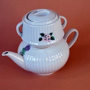 Könnte beinhalten: Eine weiße Keramik-Teekanne für eine Person mit stapelbarem Design und einem abnehmbaren Deckel, der als Tasse dient. Die Außenseite ist vertikal gerippt und sowohl an der Tasse als auch am Unterteil mit kleinen Blumenmotiven in Rosa, Grün und Violett verziert.