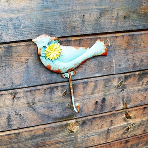 Peut inclure: Un oiseau en métal bleu avec une fleur jaune sur la poitrine est fixé à un mur en bois. L'oiseau est un crochet.