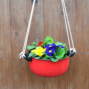 Può includere: Un vaso di metallo rosso appeso a una corda bianca, pieno di fiori colorati, tra cui gialli, blu e rossi.