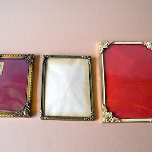 Può includere: Tre cornici per foto vintage con dettagli dorati e bronzo ornati. Le cornici rettangolari presentano uno sfondo rosso, bianco e bordeaux. Le cornici sono disposte su una superficie rosa chiaro, mostrando i loro intricati disegni angolari.