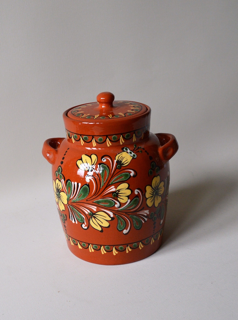 May include: A terracotta jar with a lid, decorated with hand-painted yellow flowers and green leaves on a red background.