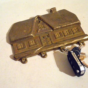 Könnte beinhalten: Ein bronzefarbener Schlüsselhalter in Hausform mit Strohdach, Fenstern und einer Tür. Ein Satz Schlüssel, einschließlich eines Autoschlüssels, hängt von unten. Der Schlüsselhalter ist aus Metall und hat ein rustikales Aussehen.