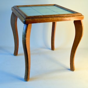 Könnte beinhalten: Kleiner, quadratischer Holztisch mit hellbraunem Finish. Die Tischplatte hat ein Rastermuster unter Glas. Der Tisch hat vier geschwungene Beine, die ihm Eleganz verleihen. Der Tisch ist etwa 61 cm hoch.