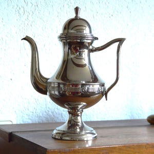 Puede incluir: Una tetera de plata con tapa abovedada y una banda decorativa alrededor del cuerpo. La tetera tiene un pico y una asa y se asienta sobre una base de pedestal.