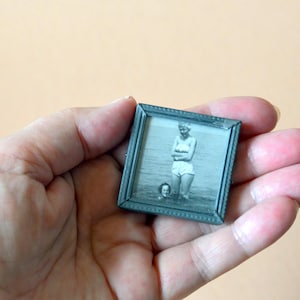Può includere: Una piccola cornice quadrata color argento che contiene una fotografia in bianco e nero di due persone nell'acqua. Una persona è in piedi, l'altra è sommersa. La cornice è tenuta in una mano aperta.
