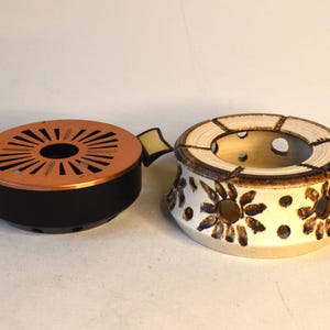 Può includere: Due pentole per fondue vintage, una con base nera e parte superiore in rame con un design circolare, e l'altra con base bianca con un design floreale marrone.