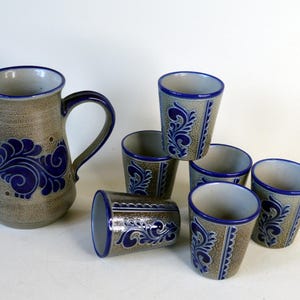 Może przedstawiać: Ceramiczny dzbanek i pięć pasujących kubków z nakrapianą beżową podstawą oraz ręcznie malowanymi kobaltowymi wzorami kwiatowych zawijasów. Każdy element ma gładki niebieski brzeg i teksturowane, ziemiste wykończenie na zewnętrznej powierzchni.