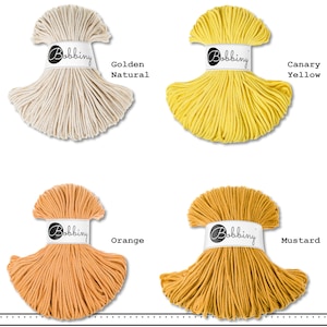 K&ouml;nnte beinhalten: Vier B&uuml;ndel Bobbiny-Kordel in einer Gitteranordnung. Die Farben sind als Golden Natural, Canary Yellow, Orange und Mustard beschriftet. Jedes B&uuml;ndel ist mit einer wei&szlig;en Papierbanderole versehen, die das Bobbiny-Logo in schwarzer Schrift zeigt.