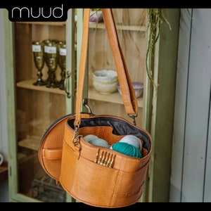 May include: A brown leather yarn bag with a long strap. The bag is open and has several balls of yarn inside, including green, blue, and white yarn. The bag is hanging on a wooden cabinet.