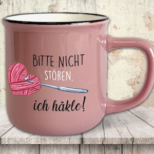 Könnte beinhalten: Eine rosa Keramiktasse mit schwarzem Rand und Henkel. Die Tasse zeigt den Text "BITTE NICHT STÖREN, ich häkle!" und ein herzförmiges Wollknäuel mit einer Häkelnadel. Die Tasse steht auf einer Holzoberfläche.