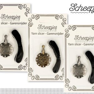 Puede incluir: Tres paquetes de cortadores de hilo Scheepjes. Cada paquete contiene un cortador de hilo plateado con un diseño decorativo y una guía de hilo negra. Los paquetes son blancos con bordes negros y el texto "Scheepjes Yarn slicer - Garensnijder" impreso en ellos.