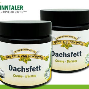 May include: Two jars of Dachsfett Creme-Balsam are shown. The jars have black lids and white labels with green and yellow accents. The text on the label includes "DAS GUTE AUS DEM INNTAL" and "gf-vertrieb.de". The jars are on a green surface.