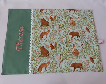 May include: A rectangular, forest-themed baby blanket. The left side is solid green with the name "Theresa" embroidered in pink. The right side features a light blue background with a pattern of brown bears, foxes, squirrels, and owls among green leaves and pink flowers.